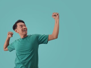 Fototapeta premium Big win. Excited Asian young man in Tosca shirt celebrating success with raised hands isolated on Tosca background, concept of celebrating victory