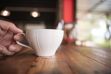 Fototapeta premium A hand holding a white cup on a rustic wooden table in a cozy café, with a blurred background and soft lighting.