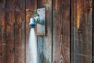 A stream of water flows under high pressure from a metal tap installed on an old wooden wall. Background.