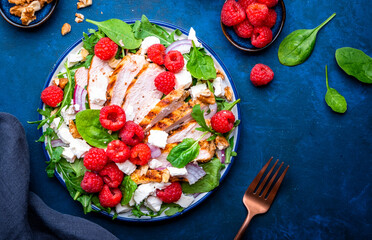 Foodie food salad with fresh raspberries, grilled chicken, feta cheese, red onion, walnuts, spinach and arugula, blue background, top view