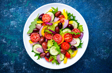 Vegan salad with olives, tomatoes, paprika, cucumber, lettuce and onion. Blue background, top view