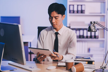 Young pensive asian man working late concentrated and serious in front of computer at night in dark...
