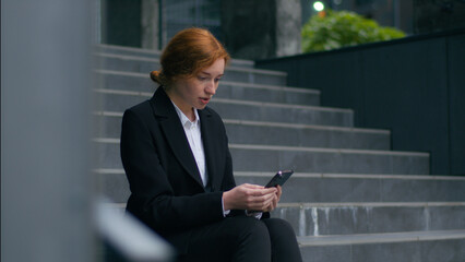 Frustrated Caucasian young businesswoman ginger girl female fired woman holding mobile phone smartphone negative message chat crying job loss bankruptcy unemployment sitting stairs outside city office