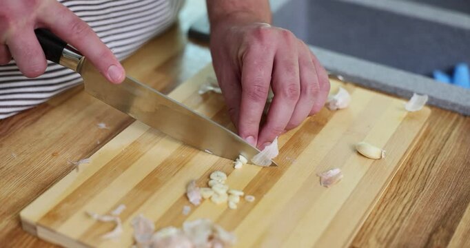 Culinarian skillfully slices peeled garlic on wooden cutting board slow motion. Preparing spicy flavorful dishes by professional chef