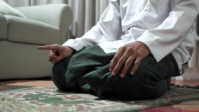 Closeup of young Asian man's hands in Muslim hat performing prayer while kneeling at home