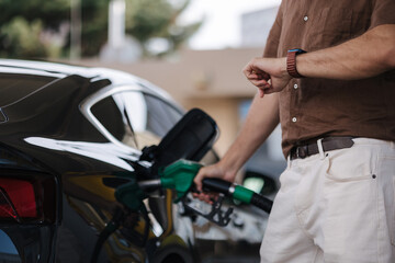 Low angle of male driver in casual clothes filling fuel into modern car with gas pump nozzle at station. Middle selection. Man looking at his watch