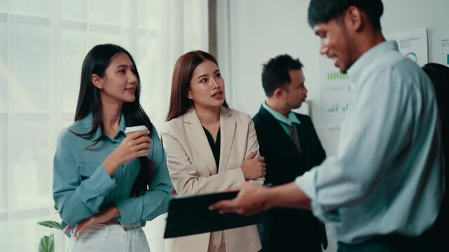 Marketing manager is showing something on his tablet to an intern during an office meeting. Other colleagues are talking in the background