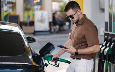 Man refueling his car on gas station and looking into his smartphone. Guy compares fuel prices in internet. Benzine. Luxury car