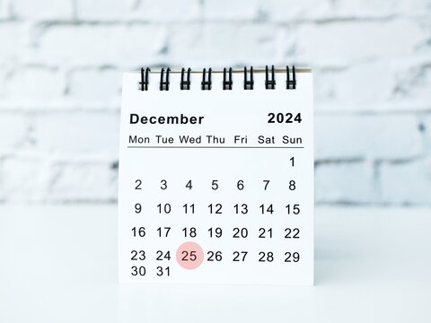 A minimalist and modern desktop calendar showing December 2025, with Christmas Day 25th highlighted by a red circle. It sits on a white table against a white brick wall