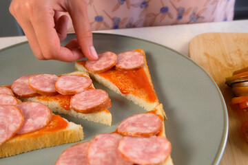 Woman putting smoked sausages slices on toasts bread with sauce cooking hot sandwiches on kitchen at home. Preparing snack fast food for herself. Tasty fresh meal for fast eating.