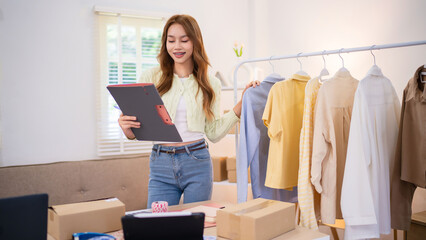 A young woman, managing her online clothing business, is holding a tablet while standing next to a clothing rack. She appears to be organizing or checking orders