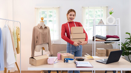 A young woman is holding several boxes while preparing orders for her online clothing business. She is smiling and working in a bright, organized workspace.