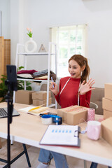 A young woman wearing a red sweater is filming content for her online clothing business. She is speaking in front of a camera, surrounded by boxes