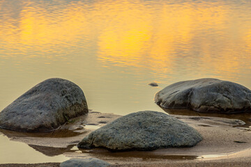 A calm beach scene with rocks scattered across the sand and a still pool of water reflecting the golden hues of a setting sun. The tranquil sea stretches into the horizon, creating a peaceful