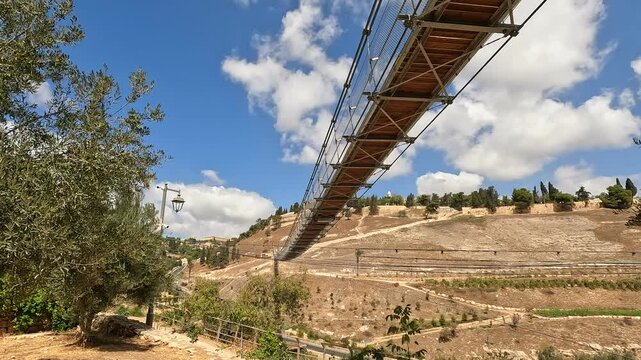 the suspension bridge over Gei Ben Hinnom near the Old City of Jerusalem, with the Mount of Olives and cloudy skies in the background, morning