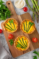 Mini tarts with green asparagus and cheese with cream filling on a wooden board on a light concrete background. Top view, selective focus.