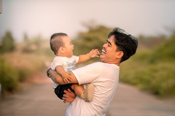 The father laughed out loud at the baby boy while lifting the baby boy. father holding baby boy outside the house in the open yard. background of natural atmosphere in the afternoon.