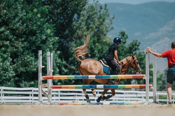 Equestrian rider and their horse jumping over a colorful obstacle during a training session outdoors with a coach.