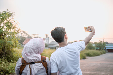 husband and wife taking a selfie together. they look happy while holding their baby on a sunny afternoon. their backs are to the camera.