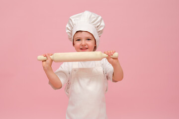 A young child wearing a chef's hat and apron holds a wooden rolling pin. Studio portrait on pink background. Kid aged 3 years (three year old boy)