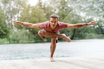 Fototapeta premium A man balances elegantly in a yoga pose on a dock, with one foot raised and arms extended. The tranquil lake and green trees provide a peaceful backdrop on a bright day.
