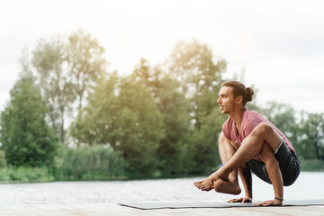 A man performs a challenging yoga pose on a mat by a serene lake as the sun sets, showcasing strength and balance amid lush greenery.