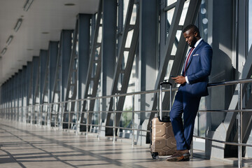 Obraz premium A man dressed in a tailored suit stands beside his wheeled luggage, focused on his mobile device while waiting in a bright, contemporary airport terminal.