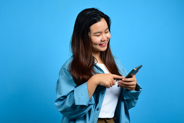 Young happy asian woman online shoping, fingers pointing while holding a cell phone in the other hand, Standing and pointing happily, light blue back ground. Copy space