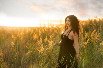 A girl with dark curly hair in a short dress among the grass on the shore of a pond at sunset. 