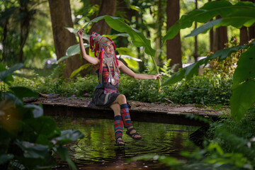 A young woman in traditional hill tribe attire is seated on a wooden bridge, holding a banana leaf in a lush green forest.