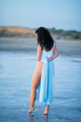 Girl with long curly hair in a bodysuit and shirt walks along the lake shore in the water at sunset.