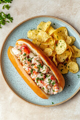 A lobster roll on a plate with potato chips, aerial view, light beige background.