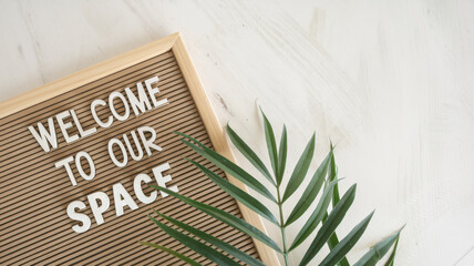 A photo of a clean background with a lot of negative space. There's a wooden board with the text "Welcome to our space". There's a green plant near the board. The background is white.