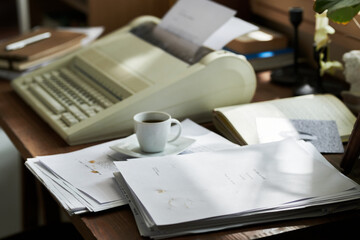 Classic typewriter sitting on wooden desk with stacks of papers and coffee cup. Scene suggests busy writing or office work environment with various documents