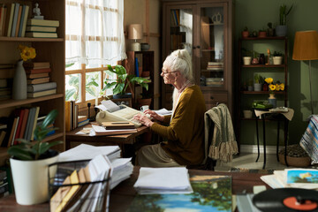 Elderly woman sitting by window reading documents in cozy room with lots of books and plants around, creating a serene atmosphere for thoughtful reflection