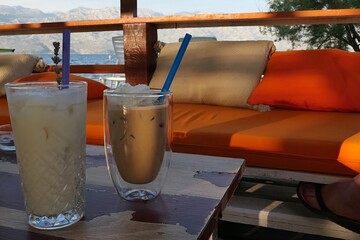 Coffee dring Latte in double wall glass and Pina Colada drink in regular glass placed on table in coastline bar, orange sofa and shoreline with Croatian Velebit mountain in background. 