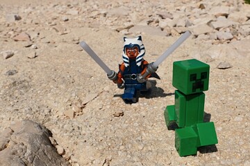 Naklejka premium ĹEGO Star Wars figure of female Jedi of Togruta race Ahsoka Tano hunting LEGO Minecraft explosive green mob with two lightsabers on sunlit summer rocky beach. 