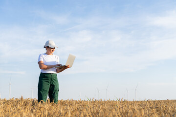 Woman farmer wearing white cap and t-shirt with laptop on agricultural field at sunset. Wind turbines in the background.. © scharfsinn86