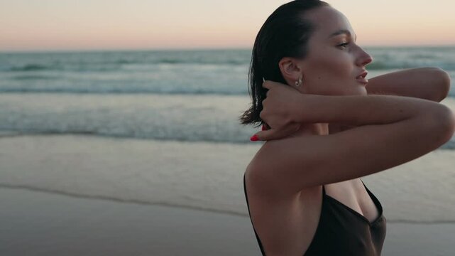 Young woman with short dark hair wearing a black swimsuit relaxes on the beach as she runs her fingers through her hair.