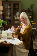 Senior woman with glasses typing on typewriter while holding crumpled paper in hand in cozy home office surrounded by bookshelves and plants