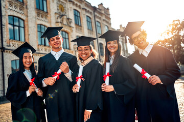  Happy smiling students in black robes and academic caps stand on the background of the university...