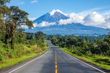 Fototapeta premium A scenic road leading towards a majestic snow-capped volcano, surrounded by lush greenery and clear blue skies.