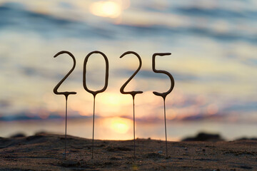 Numbers 2025 on a stick against the background of sparkling blue water and sunset on the beach. The...