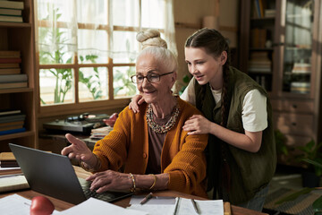 Elderly woman with white hair in bun is using laptop while young granddaughter embracing her from side in cozy room