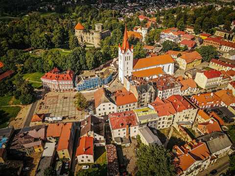 A vibrant aerial shot of a historic town, highlighting a central church with a red roof and tall steeple, alongside a medieval castle surrounded by lush green trees. The surrounding buildings feature