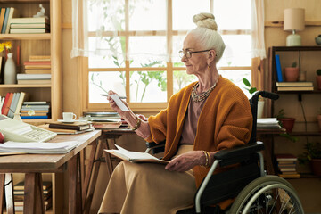 Elderly woman sitting in wheelchair writing notes at organized home office desk filled with books and potted plants on wooden shelves
