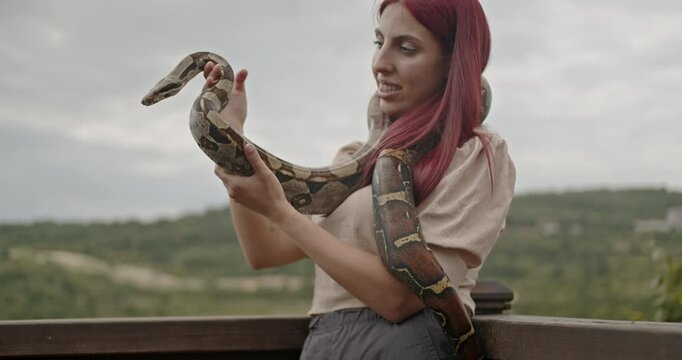 A happy young woman holds her pet snake on a balcony, smiling and laughing.