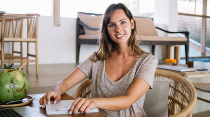 Portrait of positive female digital nomad smiling at camera while working remotely in Balinese cafe sitting at table with laptop computer and tropical coco fruit, concept of distance job in Thailand