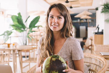 Portrait of positive cute Caucasian digital nomad holding exotic coconut beverage and smiling at...