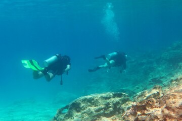 Swimming scuba divers exploring shallow ocean, rocky seabed. Scuba divers in the clear water. Underwater photography from scuba diving. Seascape with rocks, travel photo. 